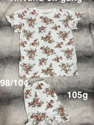 Blommig pyjamas med volangärm - Söt tvådelad pyjamas från Newbie i vitt med rosaröda blommor och gröna blad. Topp med volangärmar och matchande shorts med resår i midjan och volangkant. Perfekt för mysiga kvällar hemma.🌸 Använd en gång. Storlek 98/104 