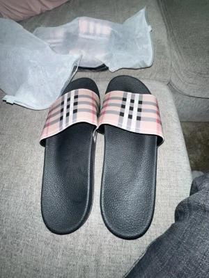 Burberry-inspirerade slides i rosa - Snygga slides med klassiskt rutmönster i rosa, svart och beige på remmen. Sulan är svart och har en lätt strukturerad yta för extra komfort. Perfekta för chill dagar hemma eller när du vill ha en trendig look.