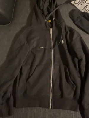 Svart hoodie från Polo Ralph Lauren - Svart hoodie med dragkedja från Polo Ralph Lauren. Klassisk design med huva och dragsko, två fickor framtill och den ikoniska broderade loggan på bröstet. Mjuk bomullskvalitet som är skön att ha på sig. Perfekt för en avslappnad och stilren look.