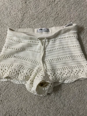 Vita virkade shorts från FB Sister - Säljer ett par vita virkade shorts från FB Sister i storlek XXS. De har snörning i midjan och ett fint hålmönster som ger en boho-känsla. Perfekta för varma dagar och riktigt sköna att bära tack vare det mjuka, stickade materialet.