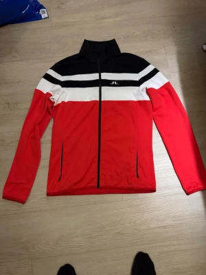 Orange och svart tröja  från J.Lindeberg - Sportig vindjacka från J.Lindeberg i orange, svart och vitt med dragkedja framtill och två sidofickor. Jackan har ståkrage, lång ärm och ett stilrent randigt mönster över bröstet. Perfekt för dig som gillar en clean och modern look.