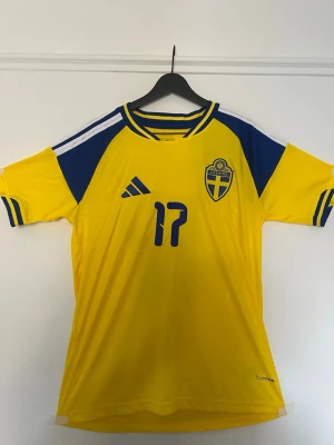 Sverige fotbollströja Adidas gul/blå - Gul och blå  fotbollströja  med Sveriges landslagsemblem på bröstet och nummer 17 tryckt framtill. Tröjan har korta ärmar, blå detaljer och klassiska Adidas-ränder på axlarna. Tillverkad i lätt och ventilerande material.