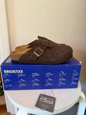 Birkenstock Boston BS tofflor mocha - Säljer ett par Birkenstock Boston BS tofflor i färgen mocha. De har ovandel i mjukt mocka, justerbart spänne i metall och klassisk korksula med mjuk fotbädd för extra komfort. Perfekta för chill dagar hemma eller ute.