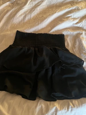 Svart volangkjol från Chelsea - Supersnygg svart kjol från Chelsea med bred smock i midjan och dubbla volanger. Kjolen har en luftig och flowy känsla, perfekt för att skapa en trendig look. Materialet är tunt och ger ett fint fall.