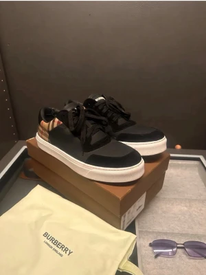 Svarta burberry sneakers - Snygga svarta sneakers från Burberry med klassiskt beige, rött och svart rutigt mönster på hälen. Skorna har vita sulor, svarta skosnören och är tillverkade i en mix av läder och textil. Perfekta för dig som vill ha en stilren men ändå unik look.