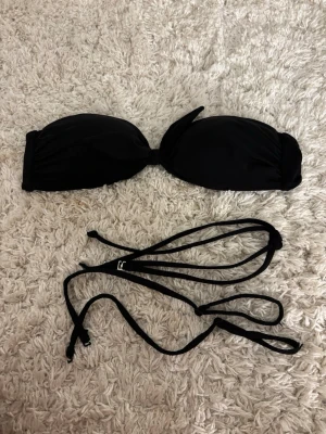 Svart bandeau bikiniöverdel - Stilren svart bandeau bikiniöverdel med avtagbara axelband och knyt i ryggen. Perfekt för dig som vill ha en enkel och klassisk look på stranden. Mjukt material som sitter bekvämt och ger bra stöd.