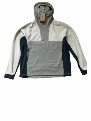 Nike ACG fleece hoodie i grått - Nike ACG hoodie i mjuk fleece med färgblock i ljusgrått, mörkgrått och svart. Tröjan har huva, dragkedjefickor på sidorna och broderad ACG-logga på bröstet. Perfekt för chill dagar eller när du vill ha en sportig look.