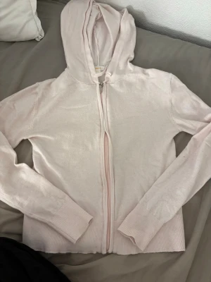 Ljusrosa hoodie från Gina Tricot - Mysig ljusrosa hoodie från Gina Tricot med dragkedja framtill och huva. Tillverkad i mjukt stickat material och har ribbade muddar vid ärmslut och nederkant. Perfekt för en chill och avslappnad stil.