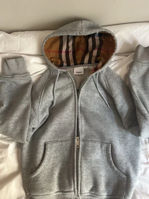 Grå zip-hoodie från Burberry - Snygg grå hoodie med dragkedja från Burberry. Klassisk huva med det ikoniska rutmönstret på insidan och broderad logga på bröstet. Mjukt material och två stora fickor framtill. Perfekt för en chill och stilren look.