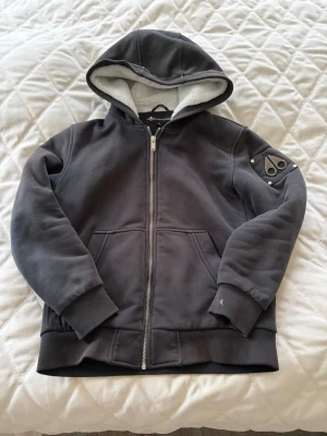 Svart hoodie från Moose Knucklers - Mysig svart hoodie med dragkedja från Moose Knucklers. Fodrad med vit teddy på insidan av huvan och quiltat foder i kroppen. Har två fickor fram och en cool logga på ärmen. Perfekt för dig som vill ha en varm och stilren hoodie med streetkänsla. Använd av 13/14 årig kille som är 168cm lång och nu är den för liten 