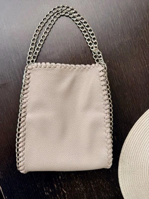 Beige handväska med kedjehandtag - Snygg beige handväska i mjukt konstläder med dubbla silverfärgade kedjehandtag. Väskan har en fyrkantig form och dekorativa kedjedetaljer längs kanterna som ger en cool och trendig look. Perfekt accessoar för att lyfta din outfit.