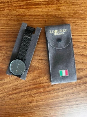 Svart klocka från Lorenzo Milano - Stilren svart klocka från Lorenzo Milano med mesh-armband i metall och minimalistisk urtavla. Urtavlan är svart med tunna vita markeringar och en liten italiensk flagga. Kommer med originalfodral.