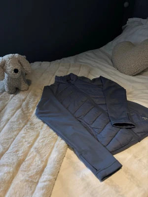 Mörkgrå pufferjacka - Snygg mörkgrå pufferjacka med släta ärmar. Jackan har en hel dragkedja och diskret logga på ena sidan. Perfekt för kalla vinterdagar och ger en sportig look.