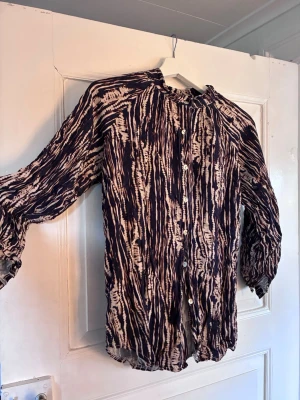 Mönstrad blus från New Collection - Snygg blus med svart och beige batikmönster från New Collection. Blusen har långa ärmar med resår vid ärmslut, knäppning framtill och en lös, luftig passform. Perfekt för dig som gillar trendiga mönster och bekväma plagg.