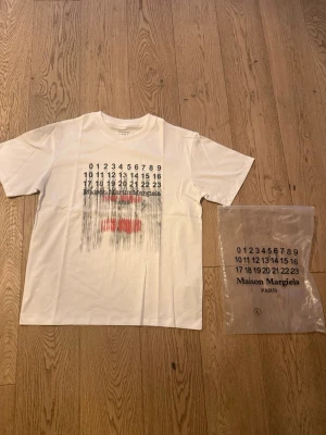Maison Margiela vit t-shirt Storlek L - Maison Margiela t-shirt i vitt med ikoniskt siffertryck och abstrakt motiv i svart och rött framtill. Lite mer baggy passform med rund hals och korta ärmar. Tillverkad i mjuk bomull, perfekt för en clean och modern look. Hör av er vid frågor pris kan diskuteras!
