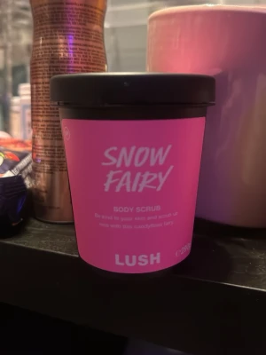 Lush Snow Fairy Body Scrub - Lush Snow Fairy Body Scrub i en rund svart plastburk med rosa etikett. Skrubben är ljusrosa och har en söt, godisliknande doft. Perfekt för att exfoliera huden och lämna den mjuk och väldoftande.