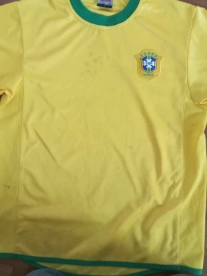 Brasilien Ronaldinho tröja gul - Gul Brasilien fotbollströja med gröna detaljer, Ronaldinho och nummer 10 på ryggen. Tröjan har rund halsringning och officiellt Brasilien-emblem på bröstet. Perfekt för fotbollsfans som vill hylla en legend. Har inte använt den super mycket den e lite stor för mig