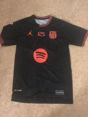 Svart FC Barcelona matchtröja Jordan - Snygg svart FC Barcelona matchtröja med röda detaljer och loggor. Tröjan har Jordan-logga, Spotify-logga och klubbmärke framtill. Tillverkad i lätt och ventilerande Dri-FIT-material, perfekt för träning eller match.