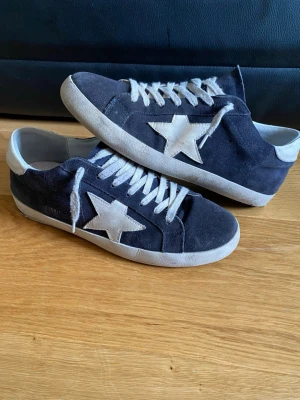 Mörkblå Golden Goose Superstar sneakers - Snygga mörkblåa sneakers från Golden Goose med vit stjärndetalj på sidan och vita skosnören. Skorna har en klassisk låg modell och är gjorda i mocka med slitna detaljer för en cool look. Perfekta för dig som gillar streetstyle och vill sticka ut.
