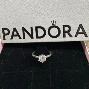 Pandora silverring med klar sten - Stilren silverring från Pandora i storlek 54, tillverkad i 925 sterling silver. Ringen har en rund, klar sten i mitten och små stenar längs bandet för extra glans. Perfekt för dig som gillar klassiska och eleganta smycken.