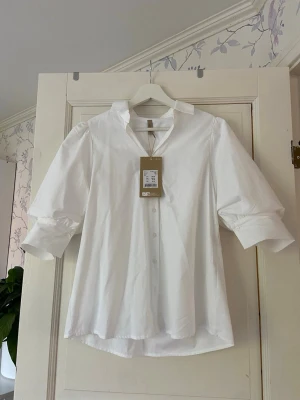 Vit skjorta med puffärm från soja concept - Stilren vit skjorta från H&M med puffiga trekvartsärmar och klassisk krage. Skjortan har knappar framtill och är tillverkad i mjuk bomull. Perfekt för en clean och fräsch look.