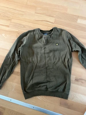 Olivgrön sweatshirt från Lyle & Scott - Olivgrön sweatshirt med rund hals och ribbade muddar från Lyle & Scott. Tröjan har det klassiska gula örnloggan broderad på bröstet. Perfekt för en avslappnad och stilren look.