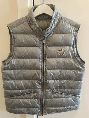 Silvergrå dunväst från Moncler - Snygg och trendig silvergrå dunväst från Moncler med klassisk logotyp på bröstet. Västen är fylld med äkta dun och har en glansig finish som ger en modern look. Perfekt för lager-på-lager-stil under kyliga dagar.