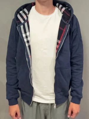 Marinblå hoodie - Tvär snygg marinblå hoodie med dragkedja och klassiskt rutigt foder i huvan. Hoodien har ribbade muddar, fickor framtill och justerbar huva med snören. Perfekt för en stilren och avslappnad look. Taggen är inte kvar, tröjan är knappt använd.