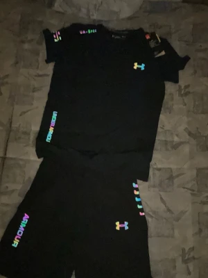 Svart träningsset från Under Armour - Snyggt svart träningsset från Under Armour med t-shirt och shorts. Båda plaggen har färgglada loggor och textdetaljer i regnbågsfärger. Perfekt för gymmet eller löprundan. Materialet känns lätt och andas bra.