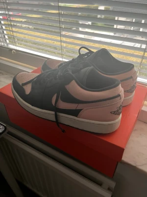 Jordan 1 Low svart/rosa/orange sneakers - Snygga Air Jordan 1 Low sneakers i svart och ljusrosa / orange läder med klassisk Swoosh på sidan och vingar på hälen. Skorna har vita sulor och svarta skosnören. Perfekta för dig som gillar streetwear och vill sticka ut med en unik färgkombo.