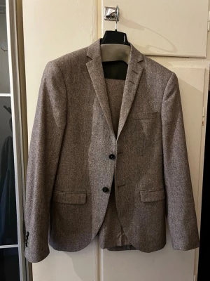Brun ullkostym med kavaj och byxor - Snygg tvådelad kostym i brun ull med klassisk kavaj och matchande byxor. Kavajen har två knappar, smala slag och fickor med lock. Byxorna har raka ben och sidfickor. Perfekt för dig som vill ha en stilren och tidlös look. Storlek 44