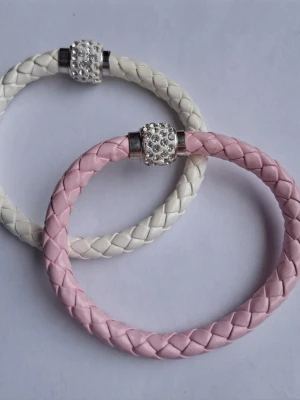 Flätade armband i rosa och vitt - Två snygga armband i flätat konstläder, ett i ljusrosa och ett i vitt. Båda har ett silvrigt magnetlås dekorerat med små pärlor som ger extra bling. Perfekta accessoarer för att lyfta din outfit.