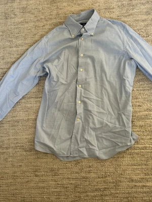 Ljusblå skjorta  - Klassisk ljusblå skjorta med lång ärm och vita knappar från Alessandro Gherardi. Skjortan har button-down krage och är tillverkad i ett tunnt, mjukt bomullstyg. Perfekt för en stilren och enkel look. Köpt från Zgander i Helsingborg!