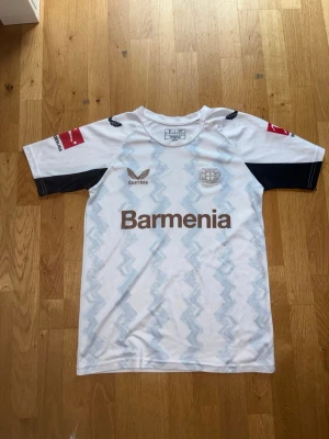 Bayer Leverkusen Xhaka matchtröja - Vit Bayer Leverkusen fotbollströja från Castore med ljusblått mönster, svarta detaljer på ärmarna och sponsorlogga Barmenia på bröstet. Nummer 34 och Xhaka på ryggen. Bundesliga-logga på ärmarna. Perfekt för fans!