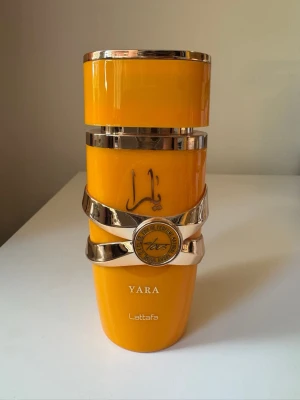 Yara parfym från Lattafa - Snygg parfymflaska i klargul färg med guldfärgade detaljer och dekorativa band. Flaskan har en modern, cylindrisk form och pryds av märket Lattafa samt texten Yara. Perfekt för dig som gillar unika och färgstarka parfymer. Ca 95% kvar. 