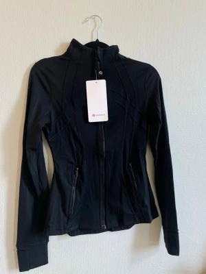 Svart define  Lululemon  - Snygg svart träningsjacka från Lululemon med figurnära passform, hel dragkedja och två fickor med dragkedja. Jackan har hög krage och tumhål vid ärmsluten. Tillverkad i ett stretchigt funktionsmaterial som andas.