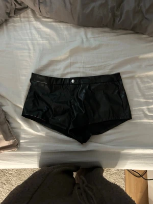 Svarta shorts i skinnimitation - Säljer ett par svarta shorts i skinnimitation med midja och knapp framtill. De har en tight passform och korta ben, perfekta för en edgy och trendig look. Passar dig som vill sticka ut och gillar att experimentera med material. Lågmidjade och så snygga 