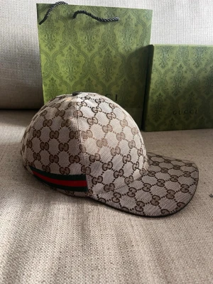 Beige Gucci Keps - Snygg keps från Gucci med klassiskt GG-mönster i beige och brunt. Kepsen har en böjd skärm och ikoniska gröna och röda ränder på sidan. Materialet är canvas och designen är riktigt trendig och lyxig.