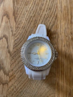 Vit klocka med strassdetaljer - Stilren vit klocka med glittrande strass runt urtavlan och vit silikonrem. Urtavlan är enkel med silverfärgade markeringar och visare. Quartz-urverk och vattentålig design. Perfekt accessoar för dig som gillar lite extra bling.