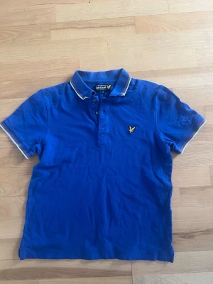 Blå pikétröja från Lyle & Scott - Snygg blå pikétröja från Lyle & Scott med korta ärmar och klassisk krage. Tröjan har broderad logga på bröstet och vita samt röda detaljer på krage och ärmslut. Perfekt för en stilren och sportig look. Medium men passar främst dig som har S, liten i strorleken.