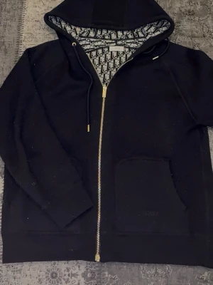 Svart hoodie med dragkedja  - Svart hoodie med guldfärgad dragkedja och huva. Insidan av huvan har ett snyggt mönster svart och vitt. Ficka framtill med diskret. Perfekt för dig som vill ha en lyxig och stilren look.