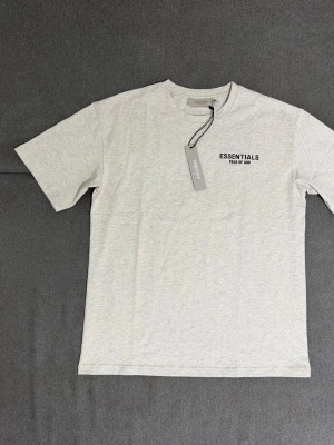 Grå Essentials Fear of God t-shirt - Snygg grå t-shirt från Essentials Fear of God med svart logga på bröstet. Klassisk rund hals och korta ärmar. Perfekt för en clean och stilren look. Tillverkad i mjuk bomull för maximal komfort.