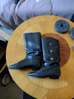 Svarta läderstövlar med spännen - Säljer ett par svarta Calsuper stövlar i äkta läder med tre dekorativa spännen i metall på ena sidan. Stövlarna har en klassisk, rak form och låg klack. Passar perfekt till både jeans och kjol för en snygg och stilren look.