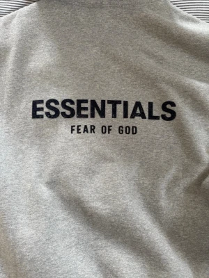 Grå Essentials Fear of God hoodie - Säljer en grå hoodie från Fear of God Essentials med svart tryck på bröstet och ryggen. Hoodien har en stor magficka, huva och ribbade muddar. Perfekt för dig som gillar streetwear och vill ha en clean look. Den är 100% äkta och är köpt från sneak