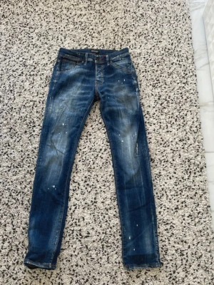 Blå jeans från Jack & Jones – modell Glen Slim 29 32 - Snygga blå slim fit jeans från Jack & Jones, modell Glenn. Jeansen har en tvättad look med slitningar och vita färgstänk för en trendig streetstyle-känsla. Klassisk femficksmodell med dragkedja och knapp. Perfekta för dig som gillar en smal passform.