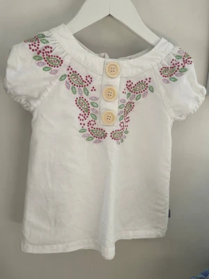Barn-Vit blus med broderi och knappar - Söt vit blus med korta puffärmar, broderade blommor i rosa, rött och grönt vid axlar och halsringning. Tre stora dekorativa knappar framtill och dragkedja bak. Perfekt för en färgglad och lekfull stil.
