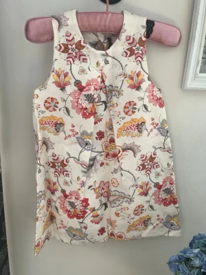 Blommig ärmlös barnkortklänning - Söt ärmlös kortklänning med färgglatt blommigt mönster i rött, gult, blått och grått på vit bakgrund. Klänningen har en enkel A-linjeform och knäppning i nacken. Perfekt för sommarens varma dagar och ger en fräsch och ungdomlig look.10 Y