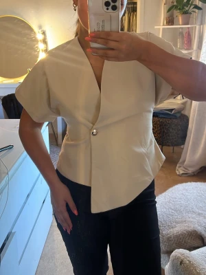 Beige omlottopp med knappdetalj - Stilren beige omlottopp med korta ärmar och asymmetrisk nederkant. Toppen har en snygg knappdetalj framtill och v-ringad hals. Perfekt för dig som gillar minimalistisk och modern stil. Köpt på Bohoo
