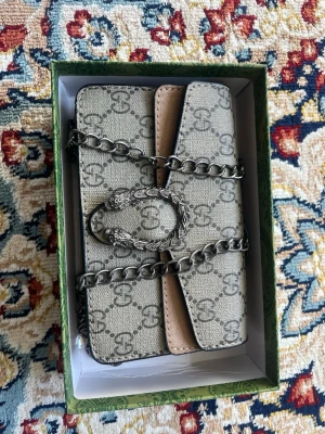 Gucci beige monogram handväska - Snygg beige handväska från Gucci med klassiskt GG-monogram över hela väskan. Väskan har en silverfärgad kedja och ett dekorativt spänne framtill. Tillverkad i canvas med detaljer i läder och metall. Perfekt för dig som gillar lyxiga accessoarer med ikonisk design.