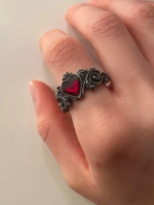 Silverring med rött hjärta - Cool ring i antiksilver med ett rött hjärta i mitten. Ringen har detaljerade snirkliga mönster och rosor på sidorna, vilket ger en vintage och romantisk vibe. Perfekt accessoar för dig som gillar unika smycken med gotisk känsla. Helt oanvänd, ej äkta 🫶